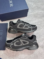 A pair of Dior low-top sneakers in black with gray and white accents, displayed next to their navy blue Dior shoebox on a gray stone floor.