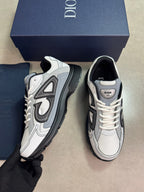 A pair of Dior sneakers in white, gray, and black, with a navy blue Dior shoebox and a navy blue dust bag in the background. A hand wearing a black glove holds one of the sneakers.