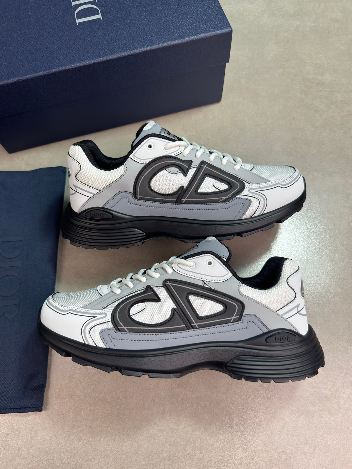 A pair of Dior low-top sneakers in white, gray, and black, displayed next to their navy blue Dior box and dust bag on a beige floor.