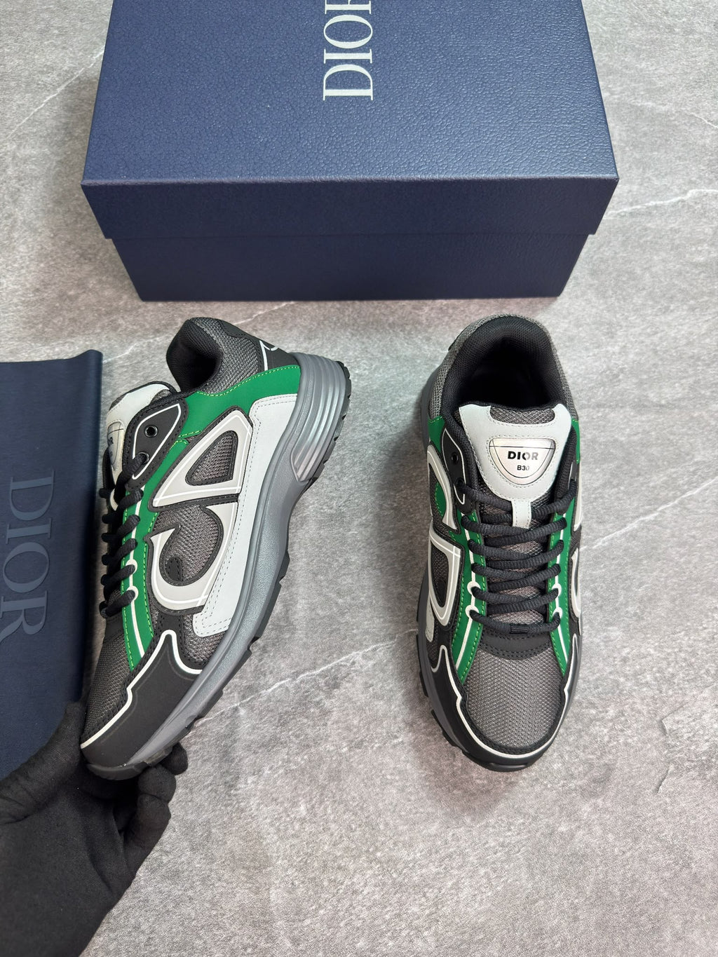 A pair of Dior sneakers in black, gray, and green, shown with their navy blue Dior box and a navy blue shoe bag on a gray stone floor. A gloved hand is holding one shoe.