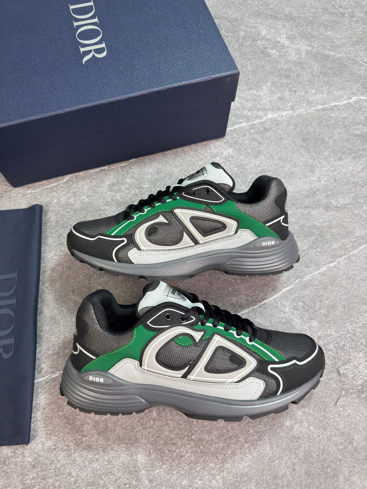 A pair of Dior low-top sneakers in black, gray, and green, displayed next to their navy blue Dior shoebox and a navy blue dust bag on a gray stone floor.
