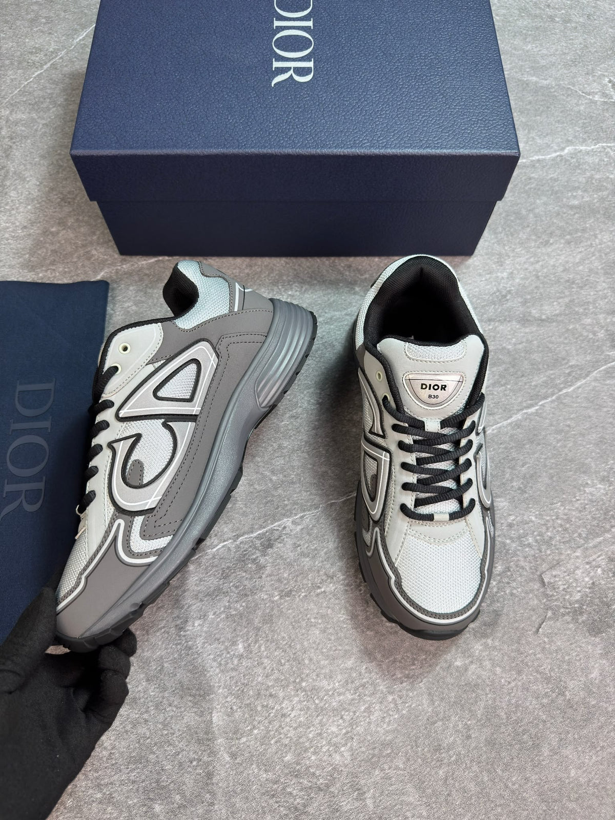 A pair of Dior low-top sneakers in gray and white with black accents, shown next to their navy blue Dior box and dust bag on a gray floor.