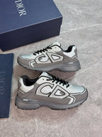 A pair of Dior low-top sneakers in gray and white with black laces, displayed next to their navy blue Dior shoebox on a gray stone-textured background.