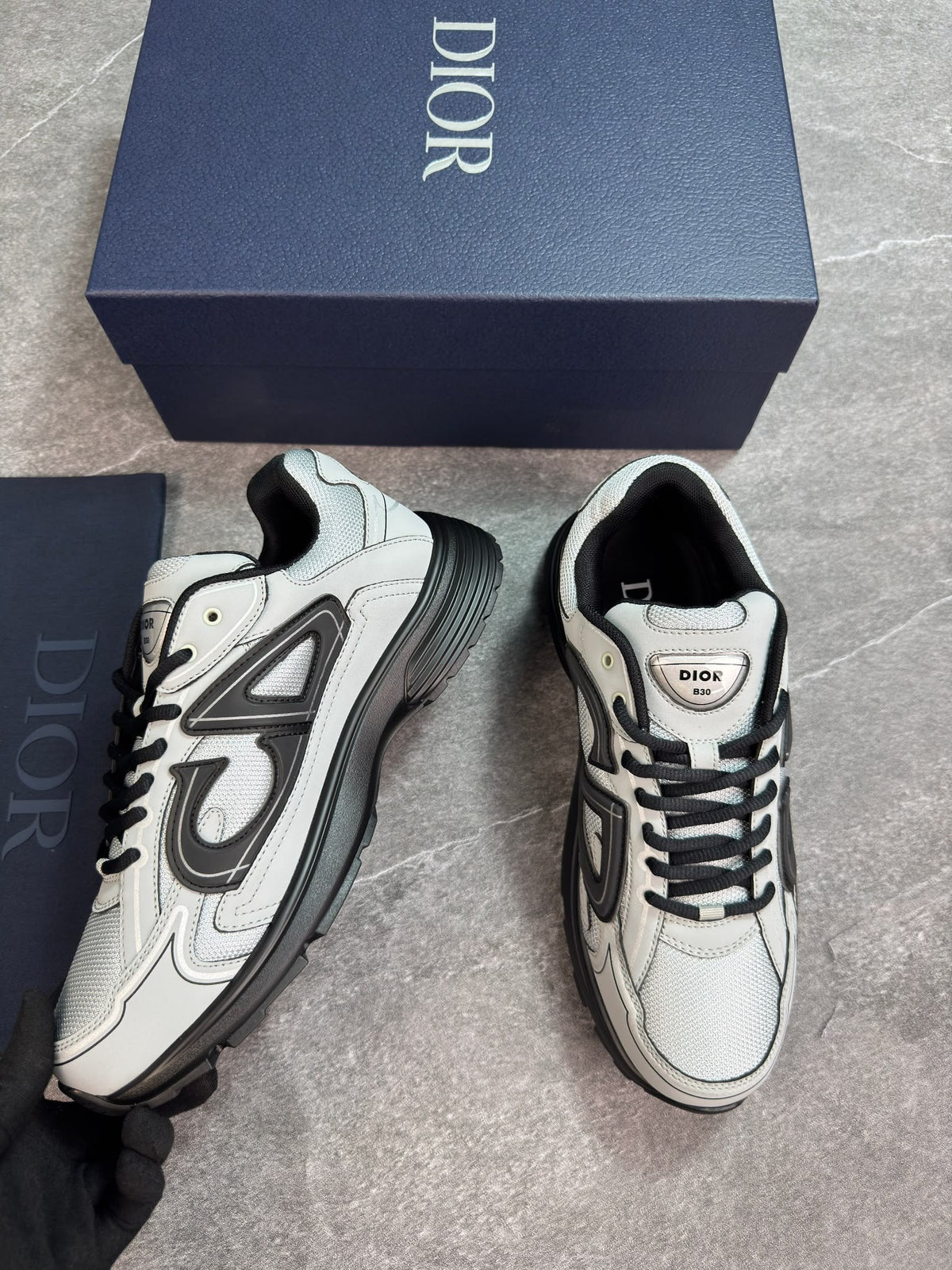 A pair of Dior sneakers in white and black, displayed next to their navy blue Dior shoebox on a gray marble floor.