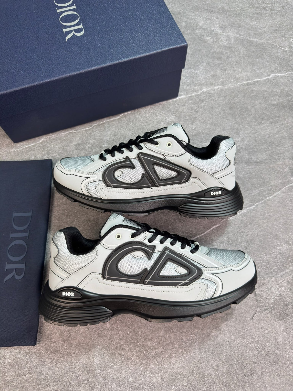 A pair of Dior low-top sneakers in white and black, displayed next to their navy blue Dior shoebox on a gray stone-textured background.