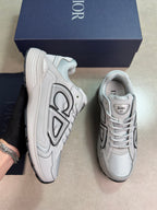 A pair of Dior sneaker low-top in light gray and white with black accents, shown next to their navy blue Dior box on a neutral beige background.