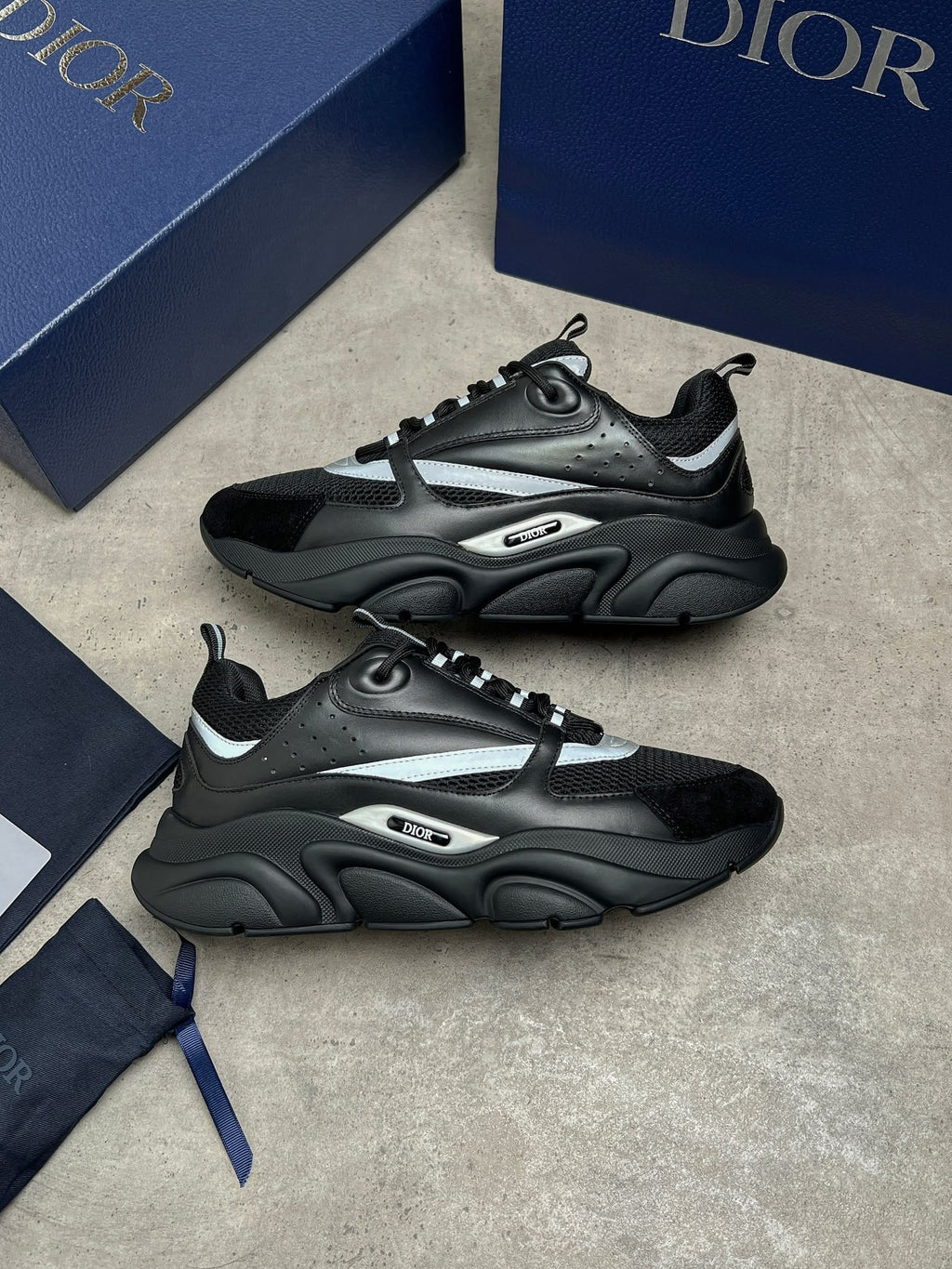 A pair of Dior low-top sneakers in black and dark gray with metallic silver accents, displayed on a gray textured surface alongside navy blue Dior shoeboxes and a dust bag.