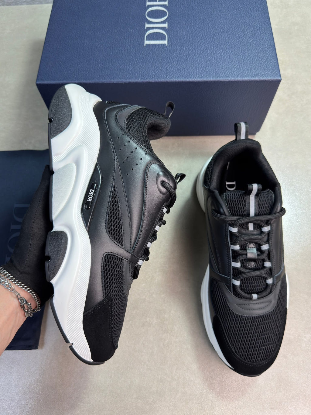 A pair of Dior low-top sneakers in black and white, with one shoe held by a hand and the other placed on a surface next to its navy blue Dior box.