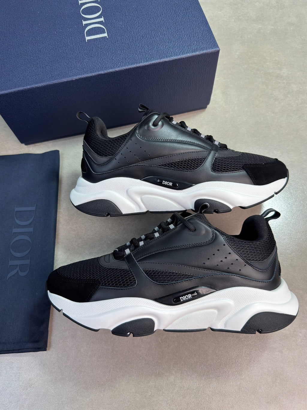 A pair of Dior low-top sneakers in black and white, displayed alongside their navy blue Dior box and dust bag on a neutral gray surface.