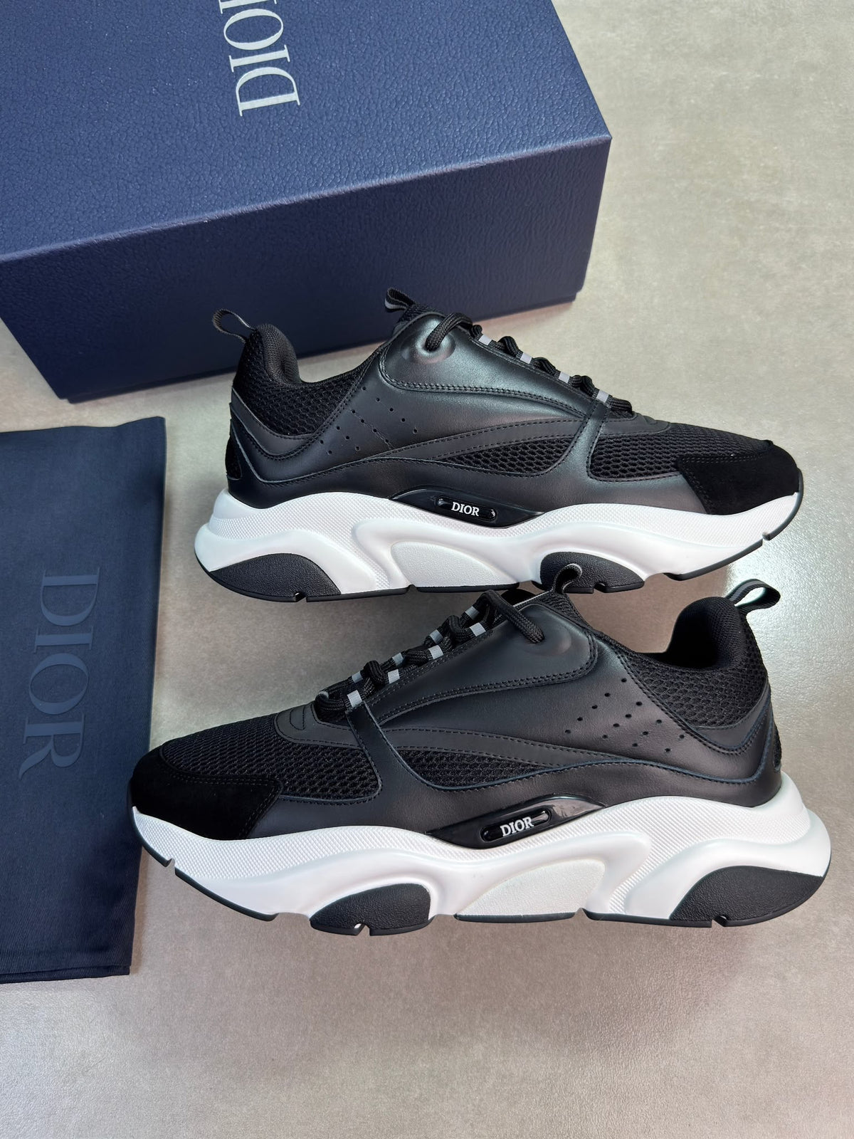 A pair of Dior low-top sneakers in black and white, displayed alongside their navy blue Dior box and dust bag on a neutral gray surface.