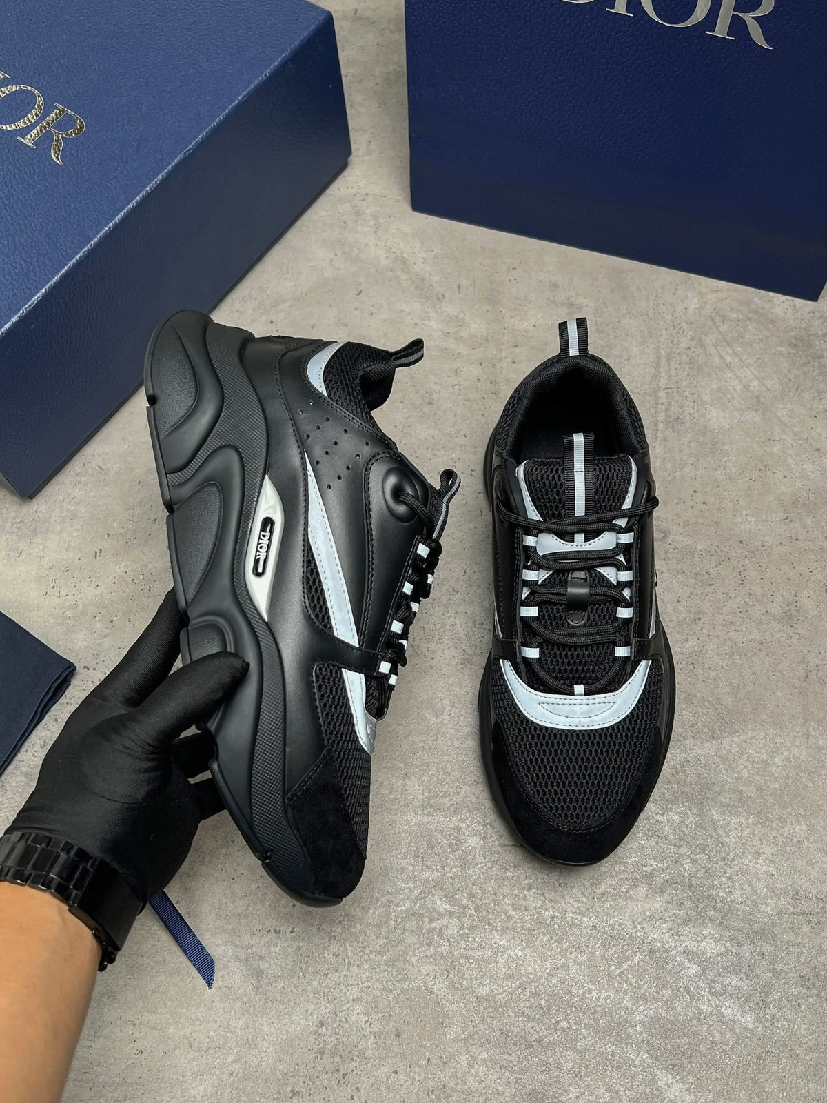 A pair of Dior sneakers in black and light gray, with one shoe held by a gloved hand and the other placed on a gray floor, with blue Dior boxes in the background.