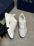 A pair of Dior low-top sneakers in off-white and cream, with one shoe held by a hand wearing a black glove and the other placed on a gray floor. Dark blue Dior shoeboxes are visible in the background.