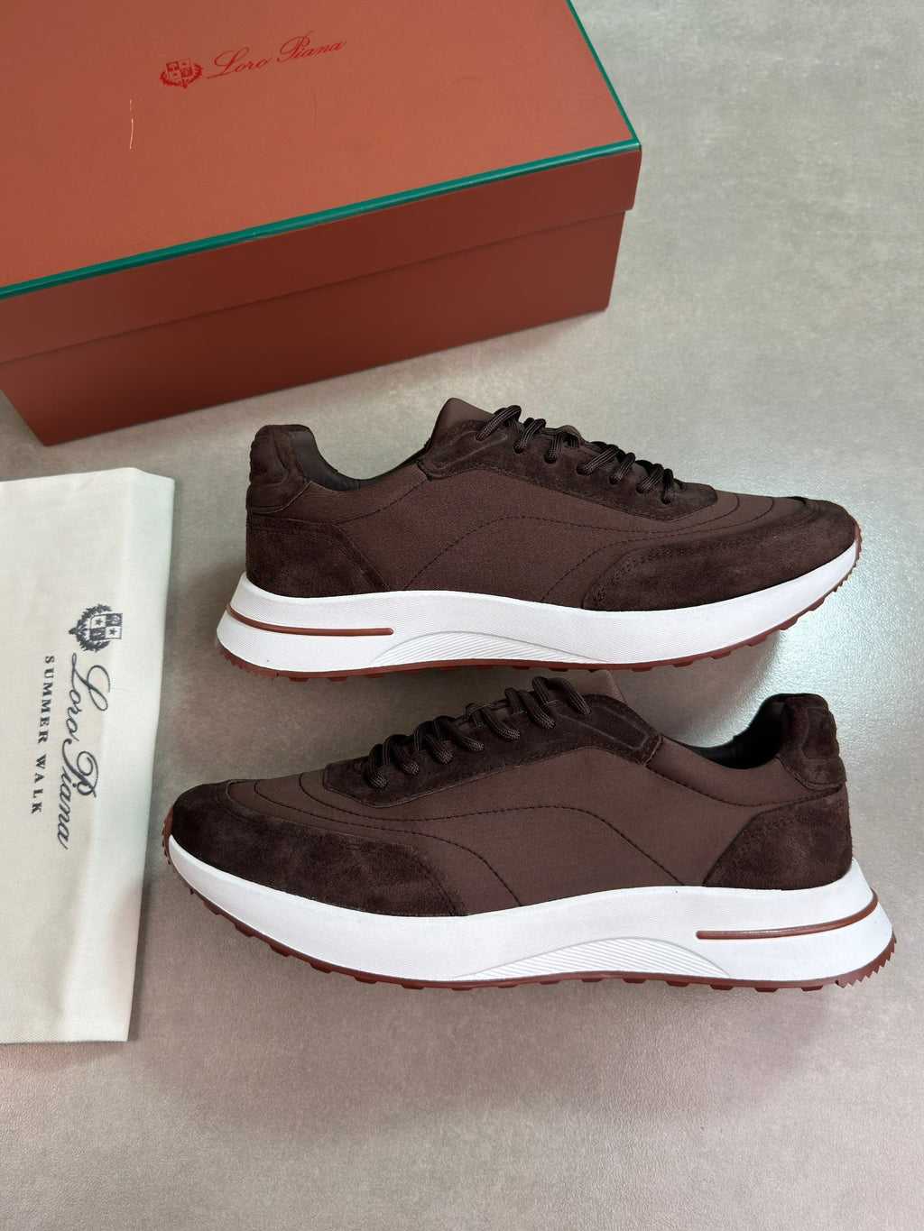 A pair of dark brown suede and leather sneakers with white midsoles and a thin red accent line, displayed next to a white branded dust bag and a brown shoebox on a gray floor.