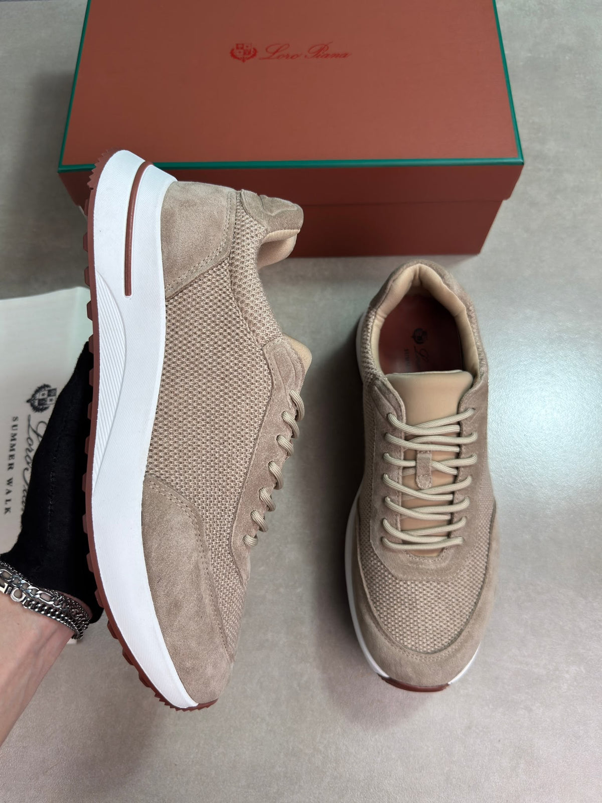 A pair of beige suede and mesh sneakers with white midsoles, shown with a brown shoebox and a branded dust bag. A hand is holding one shoe, and the other is placed on a gray floor.