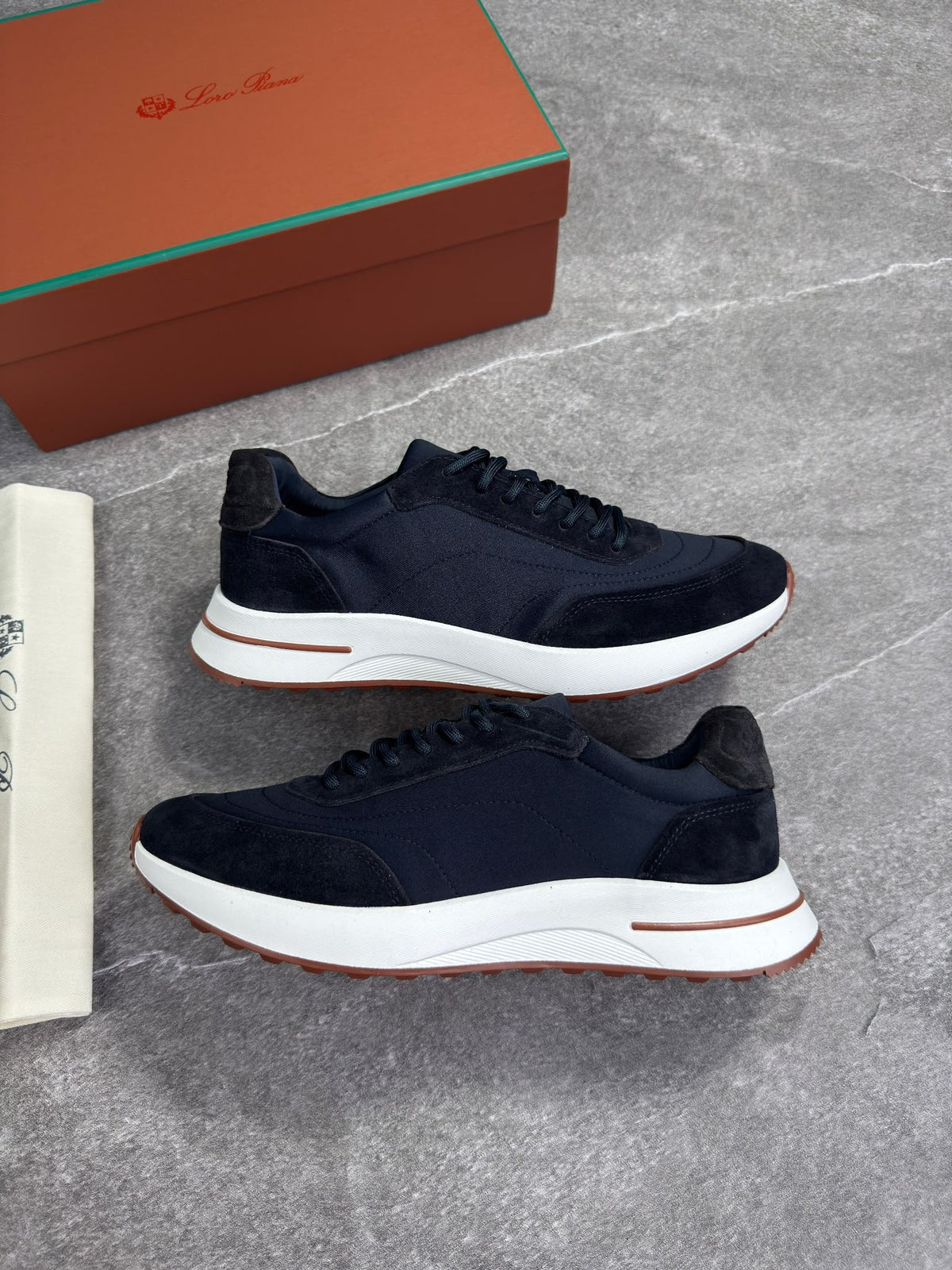 A pair of dark navy blue suede and mesh sneakers with thick white midsoles and a thin brown accent line, displayed on a gray stone-textured surface next to a brown shoebox and a white paper bag.