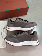 A pair of brown mesh and suede low-top sneakers with white midsoles, displayed next to a white dust bag and a brown shoebox on a gray floor.