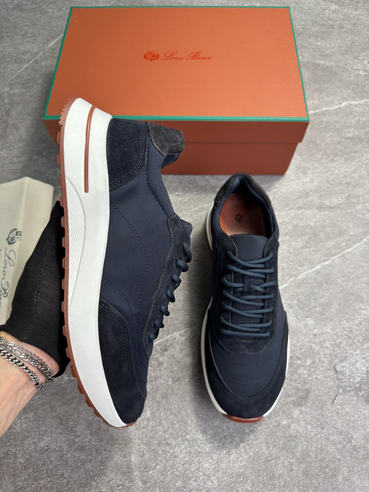 A pair of navy blue suede and canvas sneakers with white midsoles and brown outsoles, displayed next to their orange and green branded shoebox on a gray tiled floor.