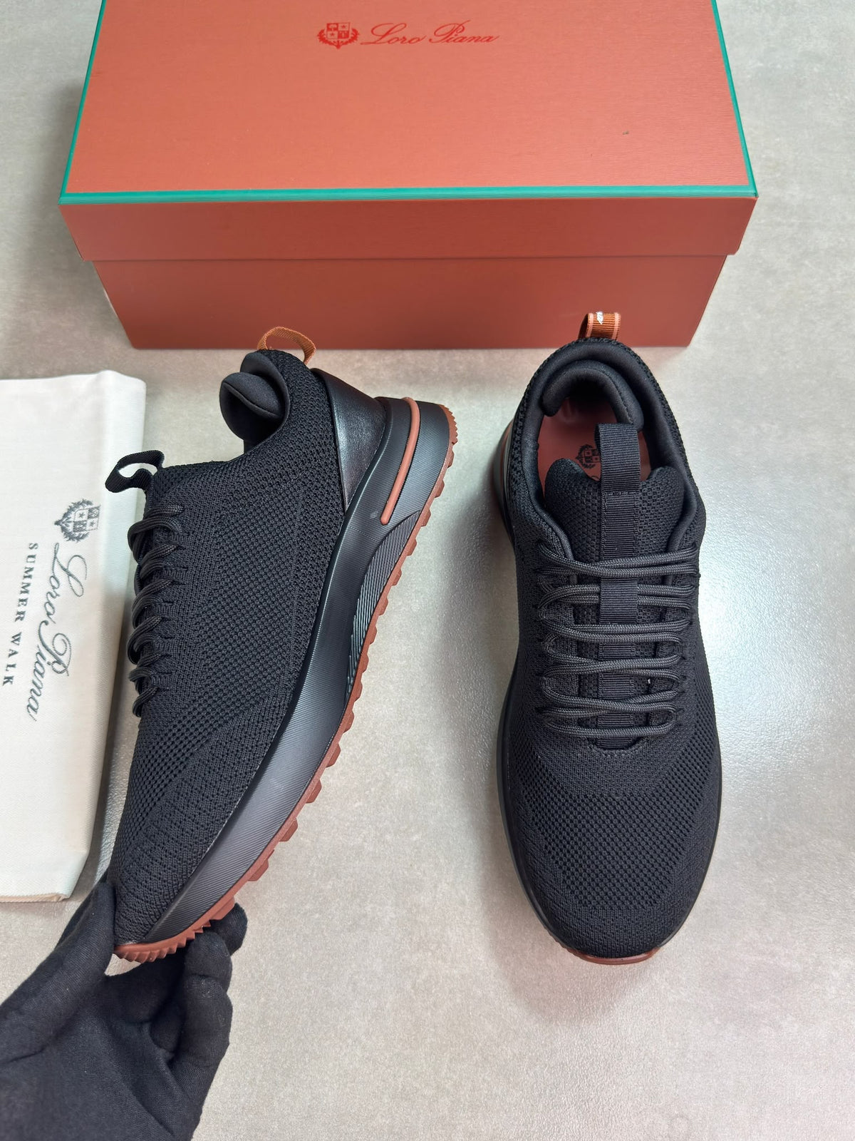 A pair of black mesh sneakers with orange-brown soles, shown with their orange and green Louis Vuitton box and a white dust bag on a light gray floor.