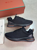 A pair of black knit athletic sneakers with dark gray soles and a thin orange-brown outsole accent, displayed on a light gray surface with a branded orange shoe box and white dust bag in the background.