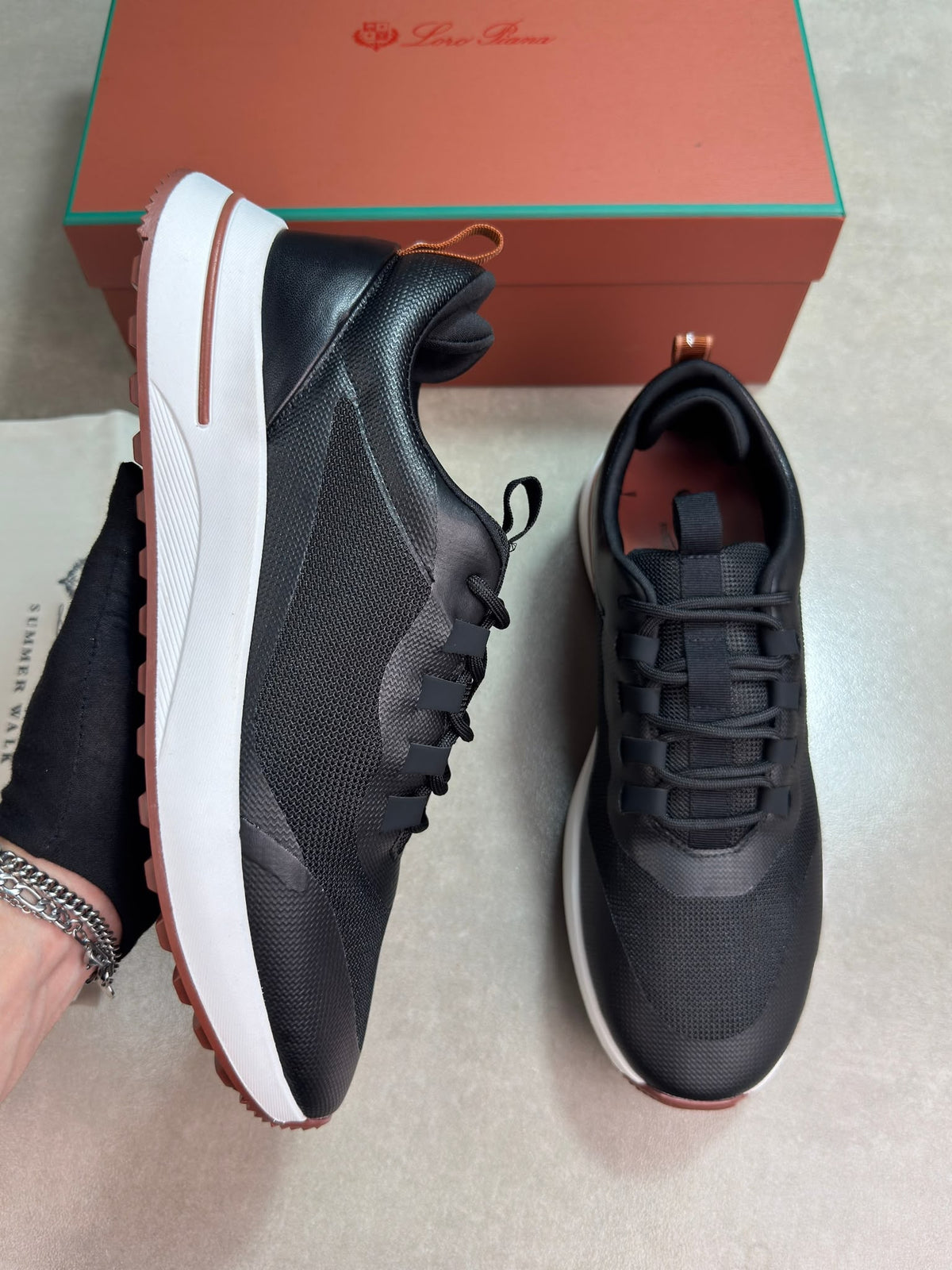 A pair of black sneakers with white midsoles and brown outsoles, shown with a branded orange shoebox in the background. A hand is holding the shoe on the left.