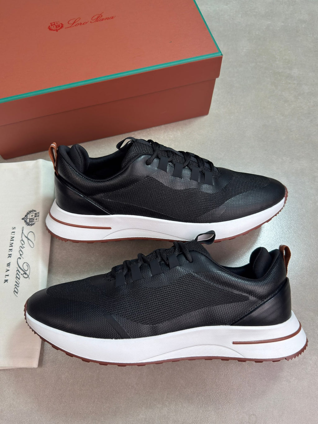 A pair of black leather and mesh sneakers with white midsoles and a thin brown outsole, displayed next to their brown and green Louis Vuitton box and dust bag on a gray floor.