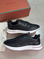 A pair of black leather and mesh sneakers with white midsoles and a thin brown outsole, displayed next to their brown and green Louis Vuitton box and dust bag on a gray floor.