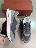 A pair of gray and white low-top sneakers with dark mesh panels and a brown pull tab, shown with a brown Louis Vuitton box in the background. One shoe is held by a hand wearing a bracelet.