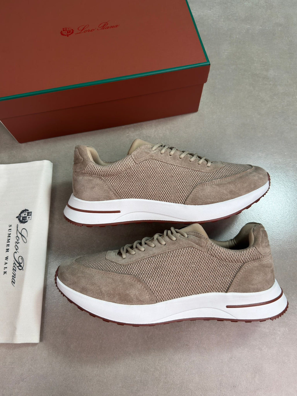 A pair of beige suede and mesh sneakers with white midsoles and brown outsoles, displayed next to a white dust bag and a brown shoebox on a gray background.