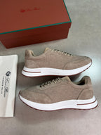 A pair of beige suede and mesh sneakers with white midsoles and brown outsoles, displayed next to a white dust bag and a brown shoebox on a gray background.