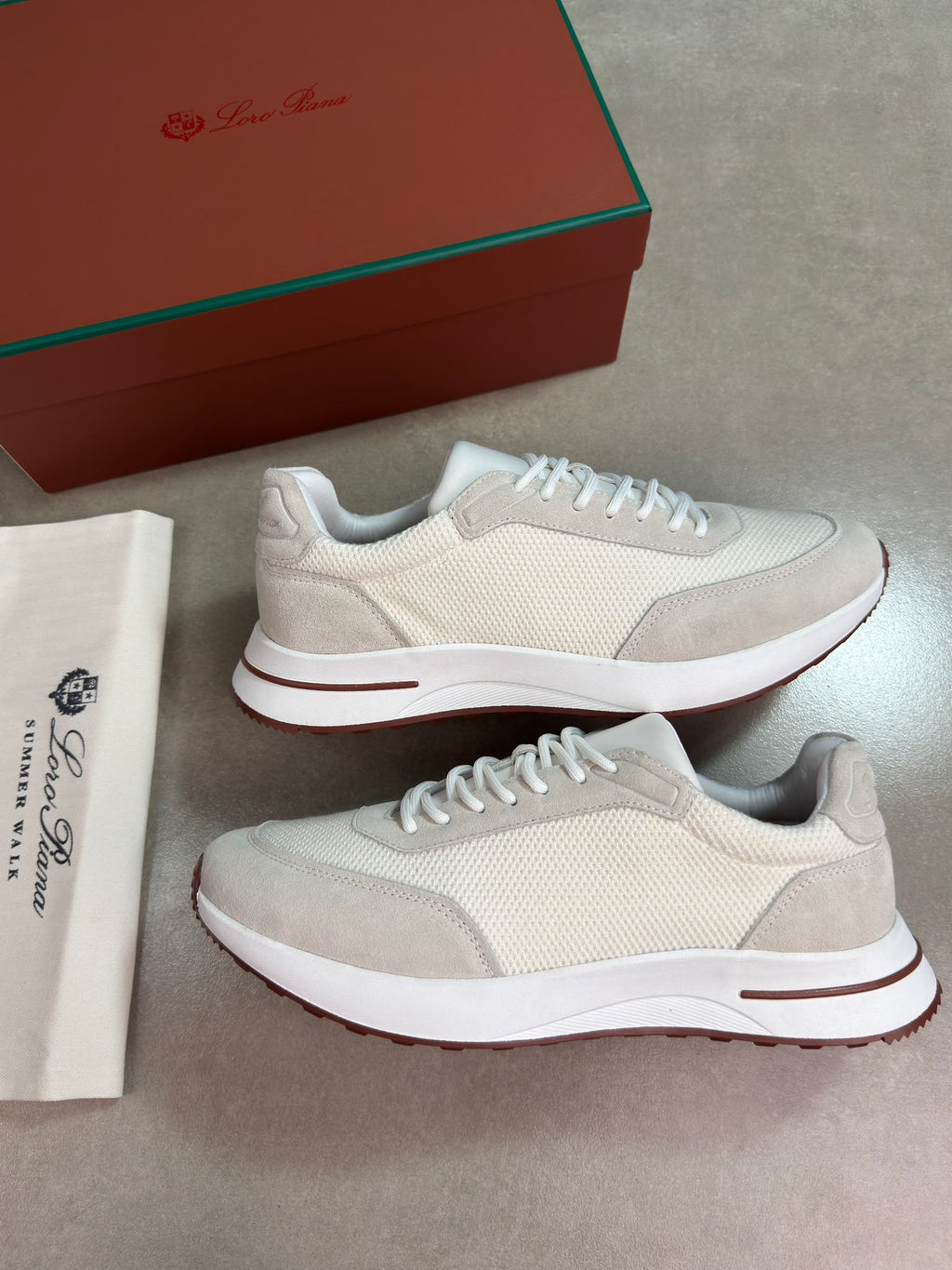 A pair of beige and white sneakers with white laces, displayed next to their brown and green branded shoebox and a white dust bag on a gray floor.