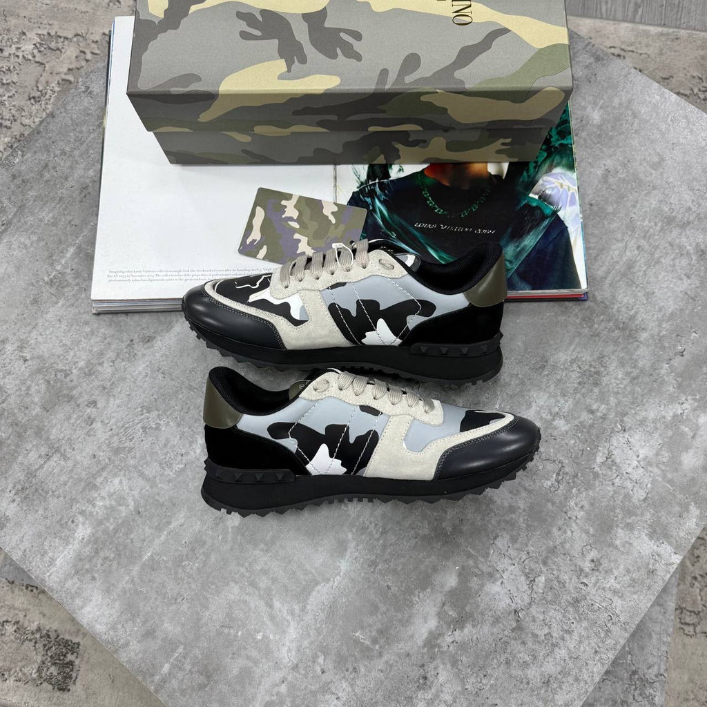 A pair of low-top sneakers with a black, white, and light gray abstract pattern, displayed on a gray stone surface with a camouflage-patterned shoebox and magazine in the background.