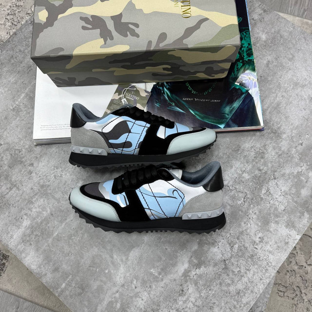 A pair of Gucci low-top sneakers in black, light blue, and gray, displayed on a gray stone surface with a camouflage-patterned shoebox and a magazine in the background.