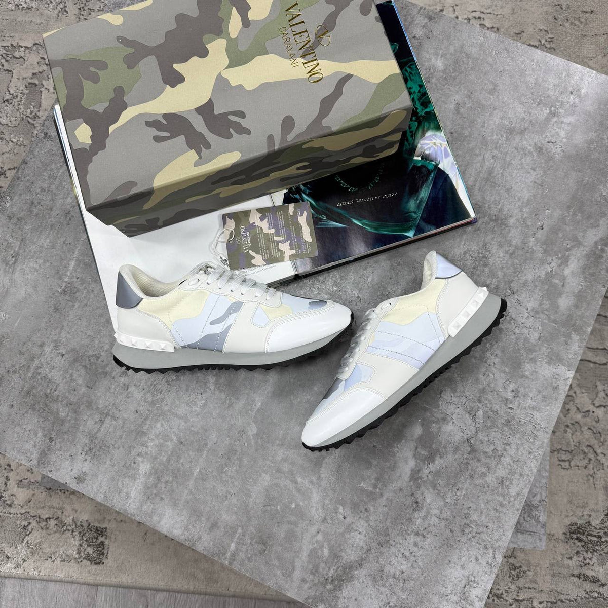 A pair of Valentino sneakers in a light cream, pale blue, and gray colorway, displayed next to their signature camouflage-patterned shoebox on a gray stone surface.