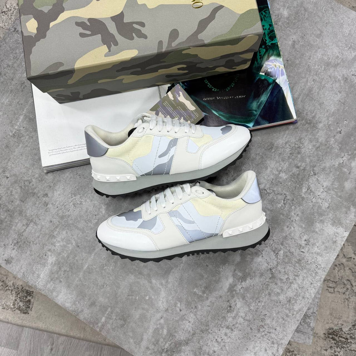 A pair of low-top sneakers in a cream, light blue, and gray camouflage pattern, displayed on a gray stone surface with a matching camouflage shoebox and a magazine in the background.