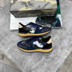 A pair of low-top sneakers with navy blue suede and black leather panels, decorated with abstract white, gray, and black brushstroke patterns, beige heel tabs, and yellow midsoles. They are displayed on a gray stone surface with a camouflage-patterned shoebox and a magazine in the background.