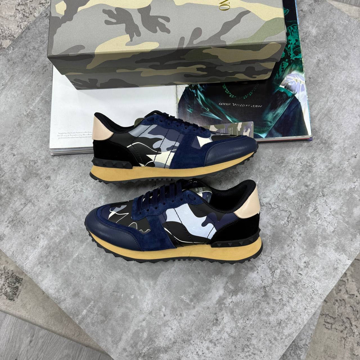 A pair of low-top sneakers with navy blue suede and black leather panels, decorated with abstract white, gray, and black brushstroke patterns, beige heel tabs, and yellow midsoles. They are displayed on a gray stone surface with a camouflage-patterned shoebox and a magazine in the background.