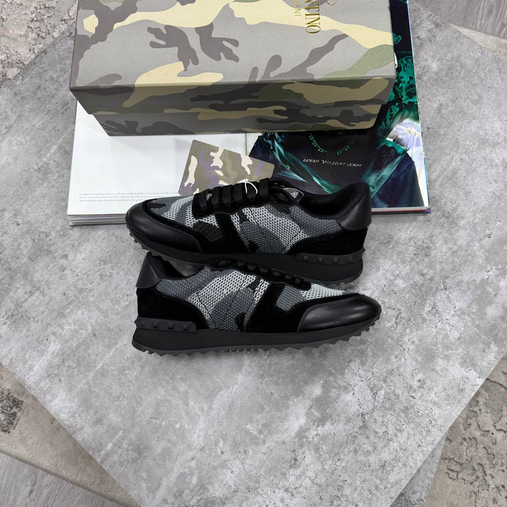 A pair of black and gray camouflage knit sneakers with black leather overlays, displayed on a gray stone surface with a camouflage-patterned shoebox and a magazine in the background.