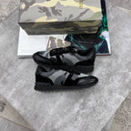 A pair of black and gray camouflage knit sneakers with black leather overlays, displayed on a gray stone surface with a camouflage-patterned shoebox and a magazine in the background.