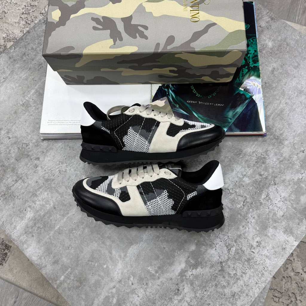 A pair of Valentino sneaker sneakers with a black and off-white patterned design, displayed on a gray stone surface with their branded box and a magazine in the background.