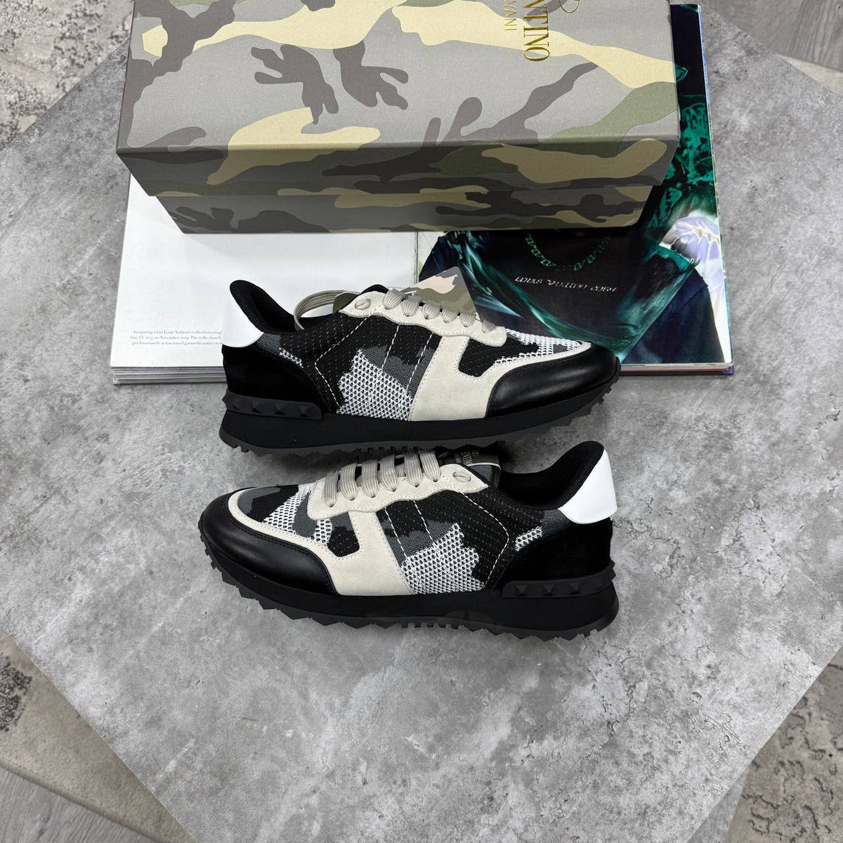 A pair of Valentino sneaker sneakers with a black and off-white patterned design, displayed on a gray stone surface with their branded box and a magazine in the background.