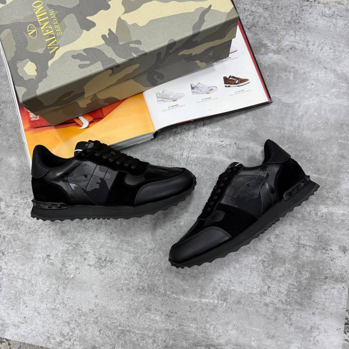 Black sneakers on a concrete surface with a camouflage shoe box in the background.