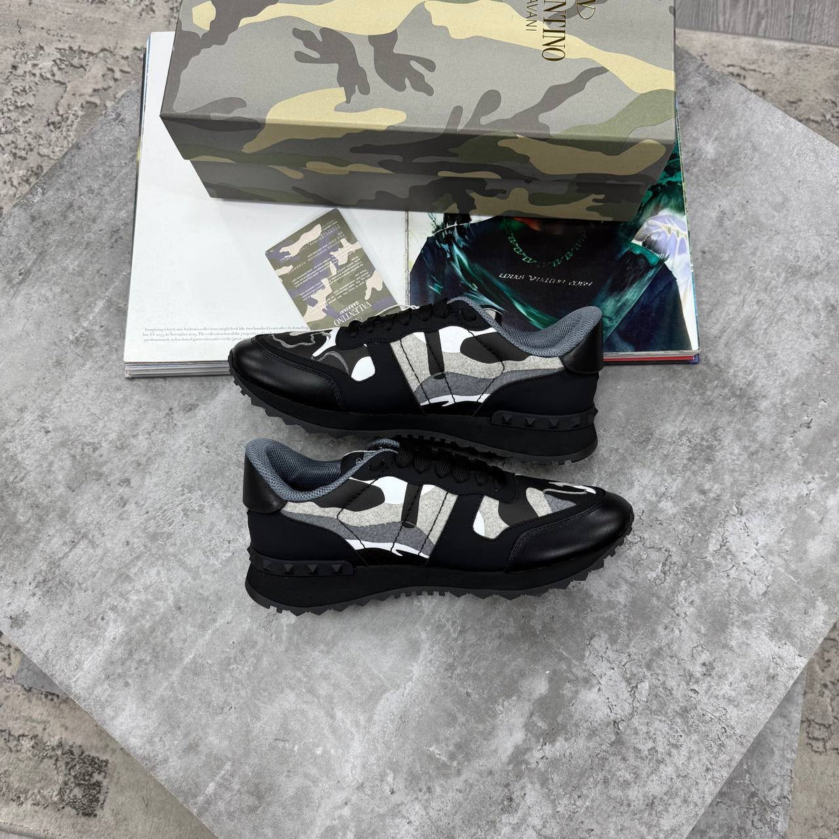 A pair of black and gray low-top sneakers displayed on a gray stone surface, with a camouflage-patterned shoebox and magazine pages behind them. The shoebox shows the brand name Valentino.