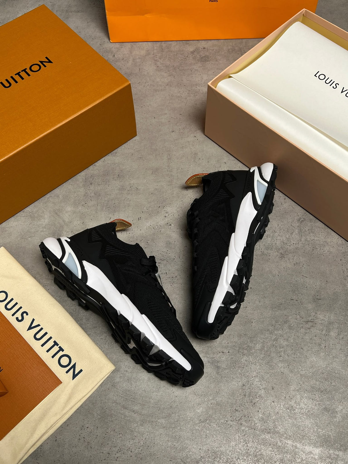 A pair of black and white Louis Vuitton sneakers displayed on a gray concrete floor alongside their signature orange and beige Louis Vuitton packaging boxes and dust bags.