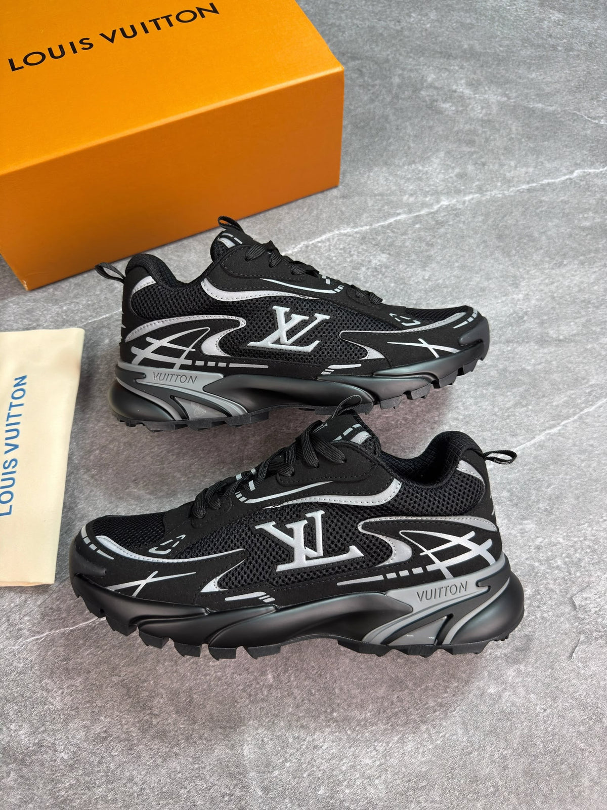 A pair of black Louis Vuitton sneakers with silver and white graphic accents, displayed on a gray textured surface next to an orange Louis Vuitton box and a beige dust bag.