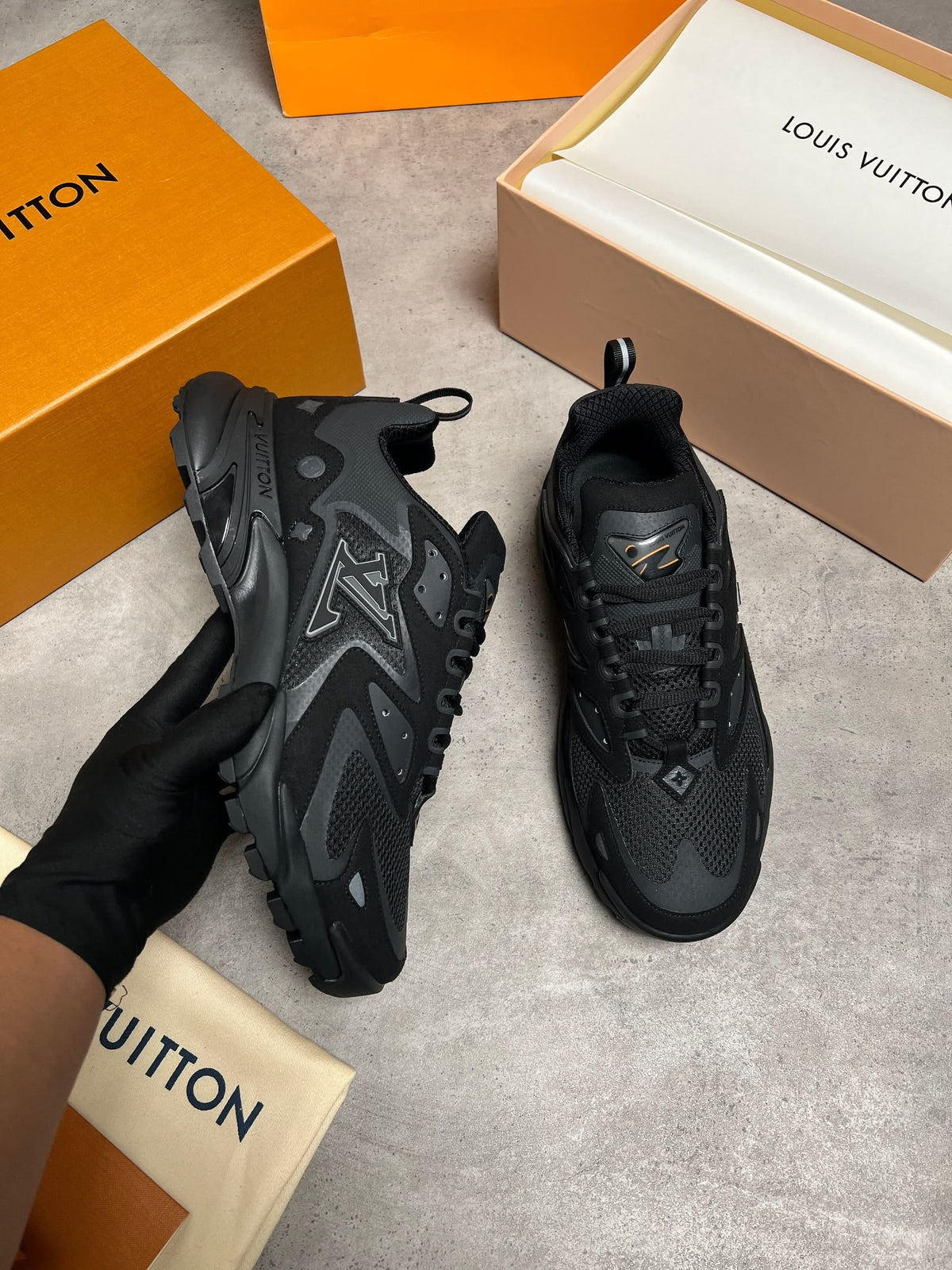 A pair of black Louis Vuitton sneakers, with one held by a gloved hand and the other placed on a gray floor, surrounded by orange and beige Louis Vuitton packaging boxes.