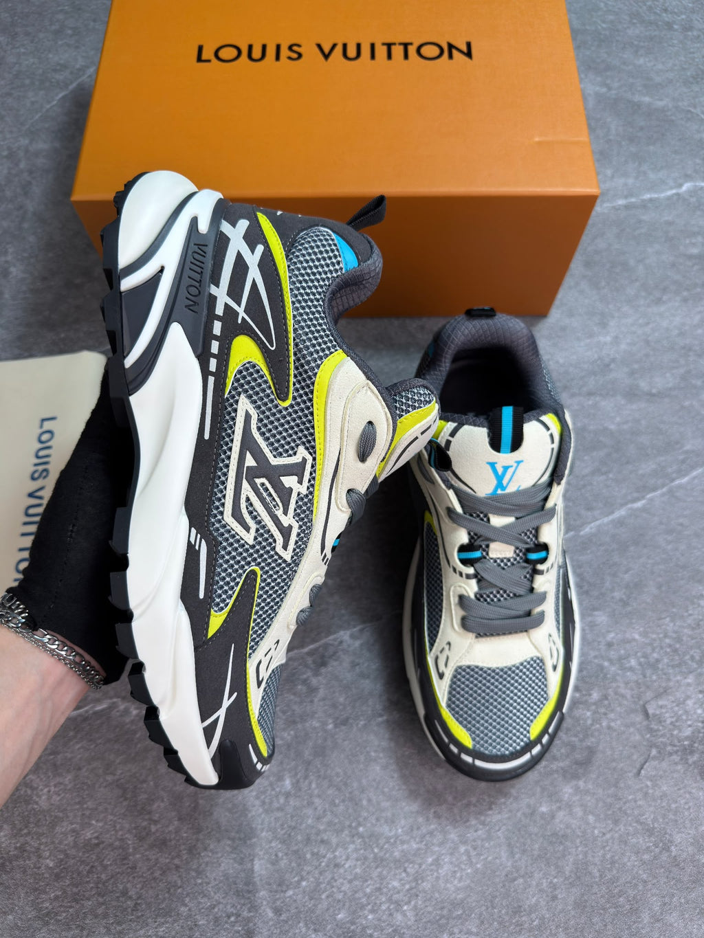 A pair of Louis Vuitton chunky sneakers in gray, white, black, and neon yellow, shown with the brand's orange box and dust bag on a gray tiled floor.
