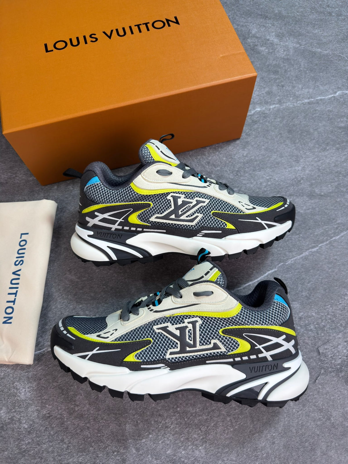 A pair of Louis Vuitton low-top sneakers in gray, black, and white with neon yellow accents, displayed next to their signature orange Louis Vuitton box and a beige dust bag on a gray textured surface.