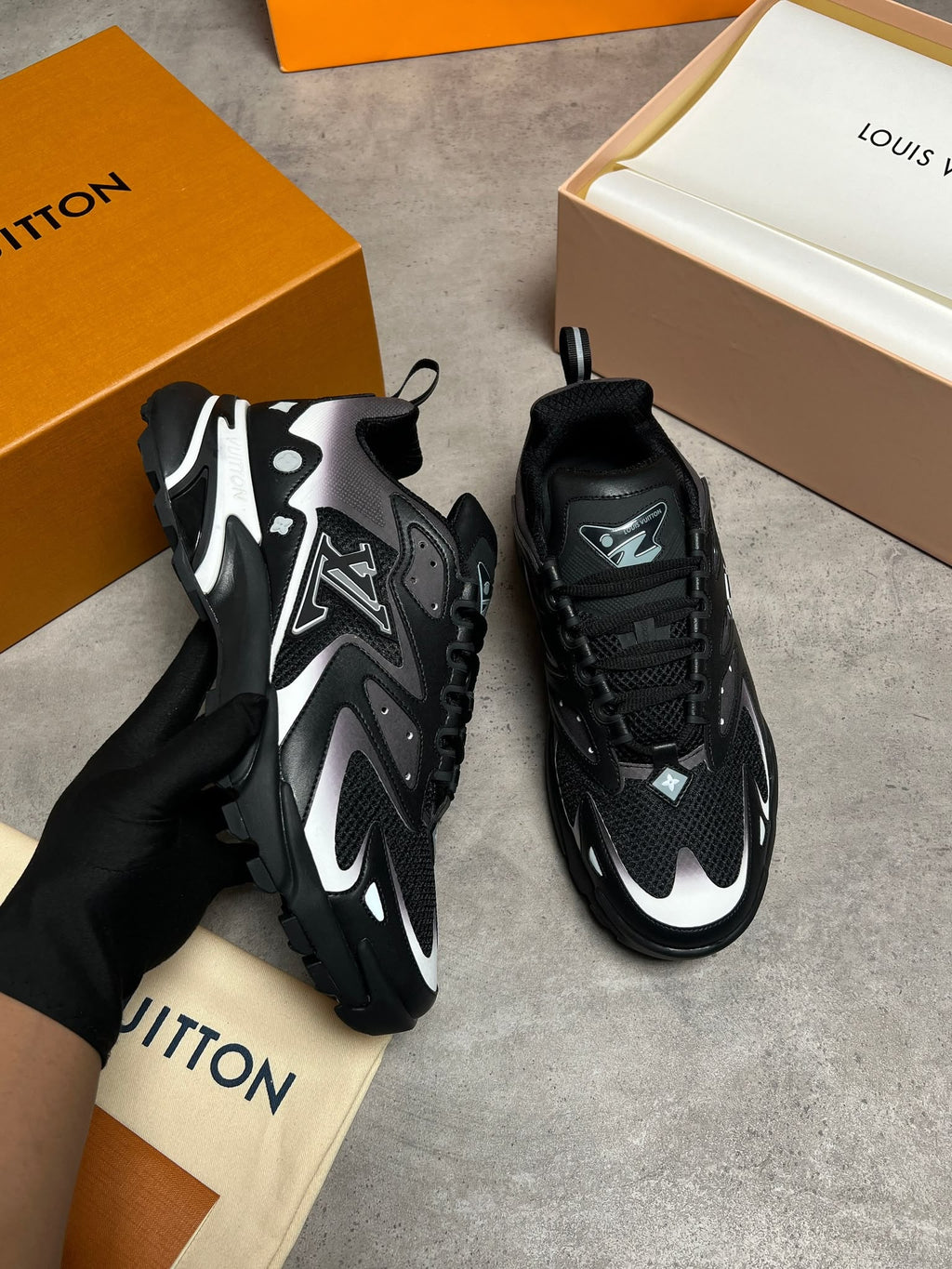 A pair of black and silver Louis Vuitton sneakers shown with branded orange boxes and a dust bag. One shoe is held by a hand wearing a black glove, while the other sits on a gray floor.