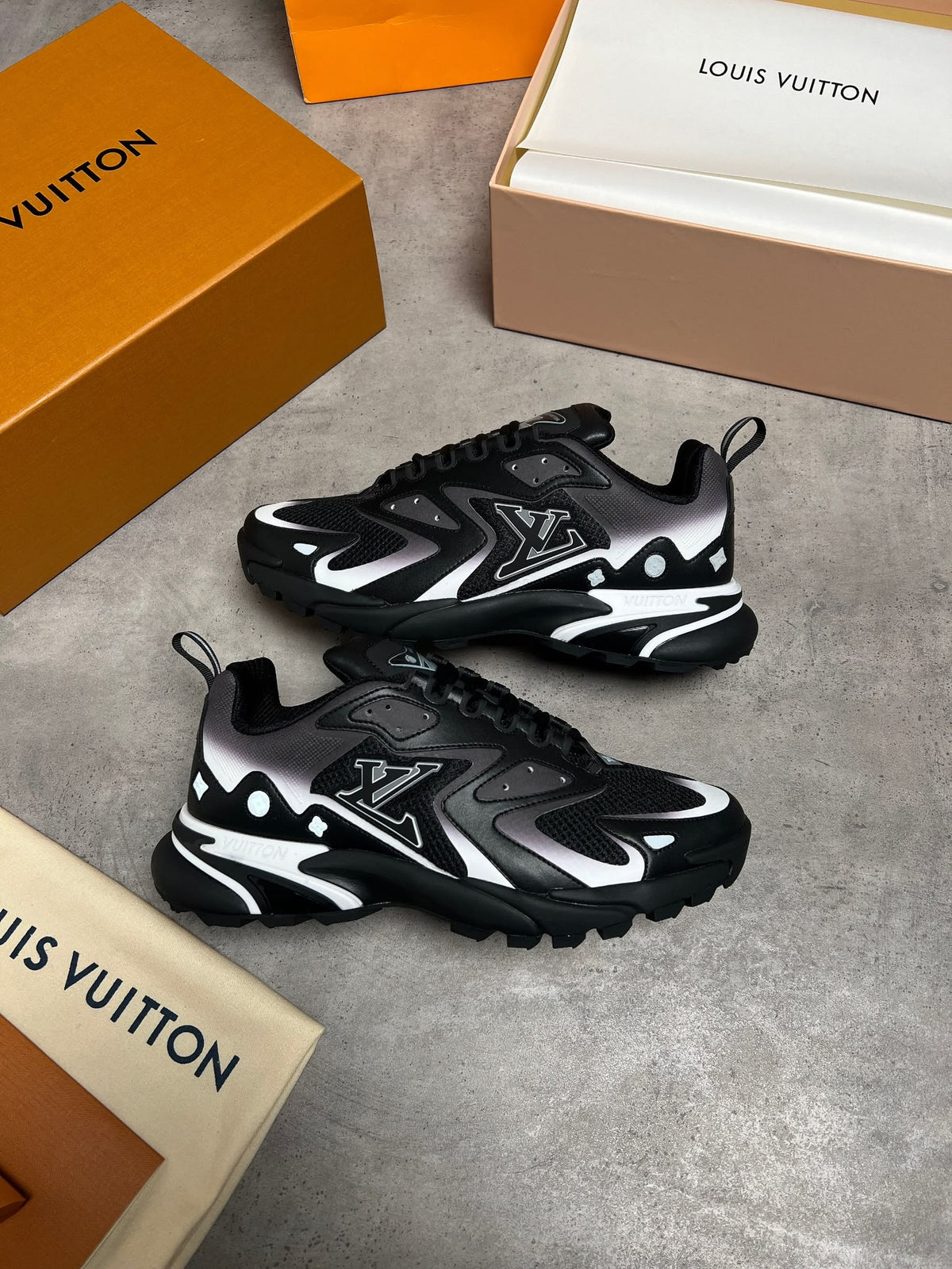 A pair of Louis Vuitton low-top sneakers in black, gray, and white, styled with branded orange and beige shoeboxes on a gray concrete floor.