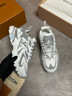 A pair of white and gray Louis Vuitton chunky sneakers, with one held by a black-gloved hand and the other placed on a gray floor. The orange and beige Louis Vuitton boxes and dust bag are visible in the background.